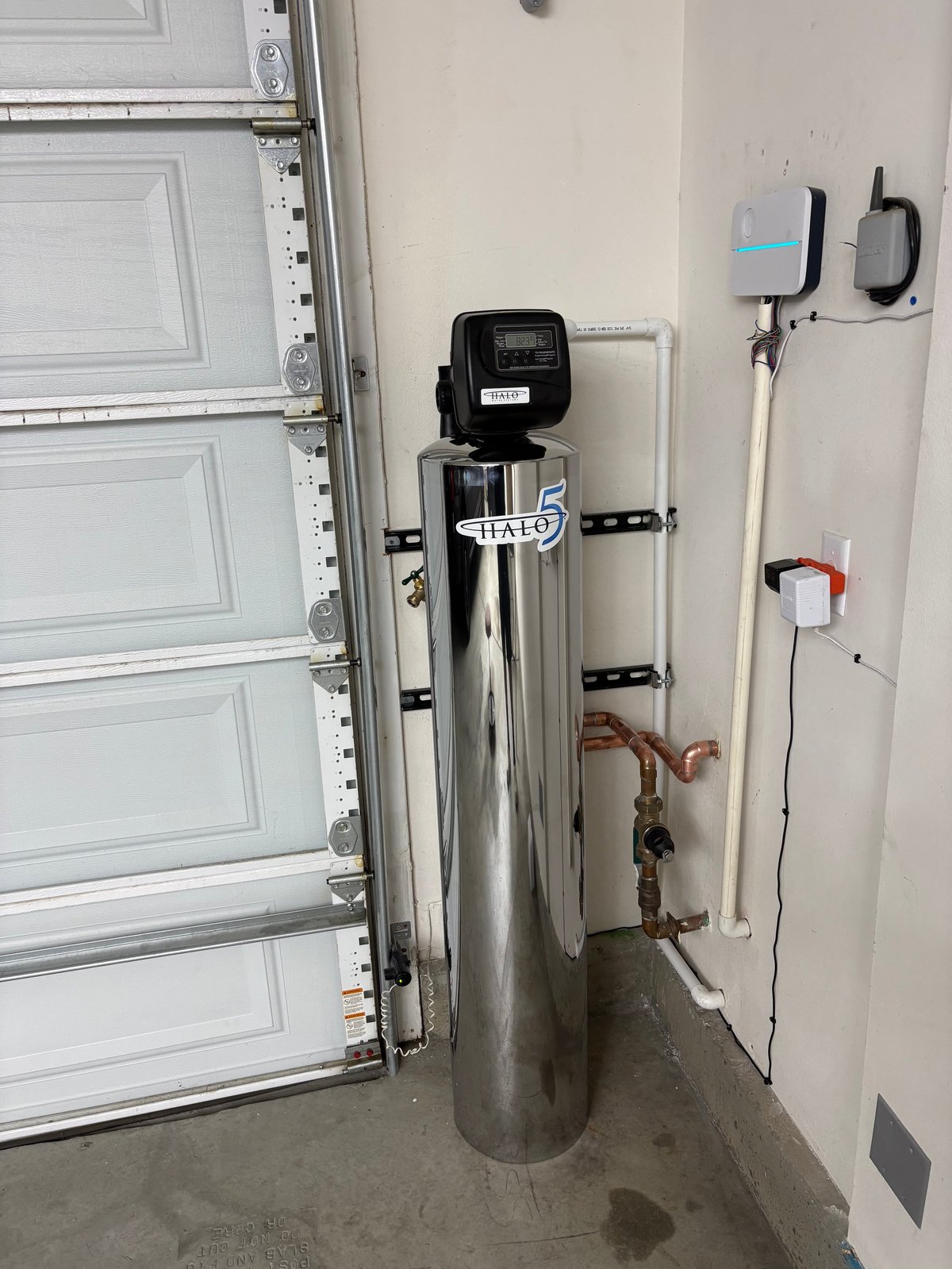 Water softener system installed in garage next to white garage door with mounted control box and pipes