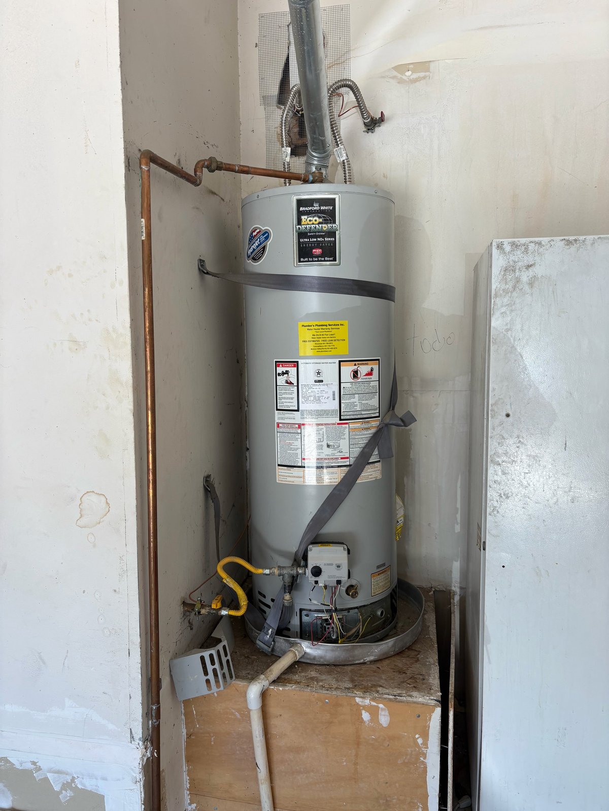 A gray water heater with copper piping installed in a utility closet with white walls and a concrete floor