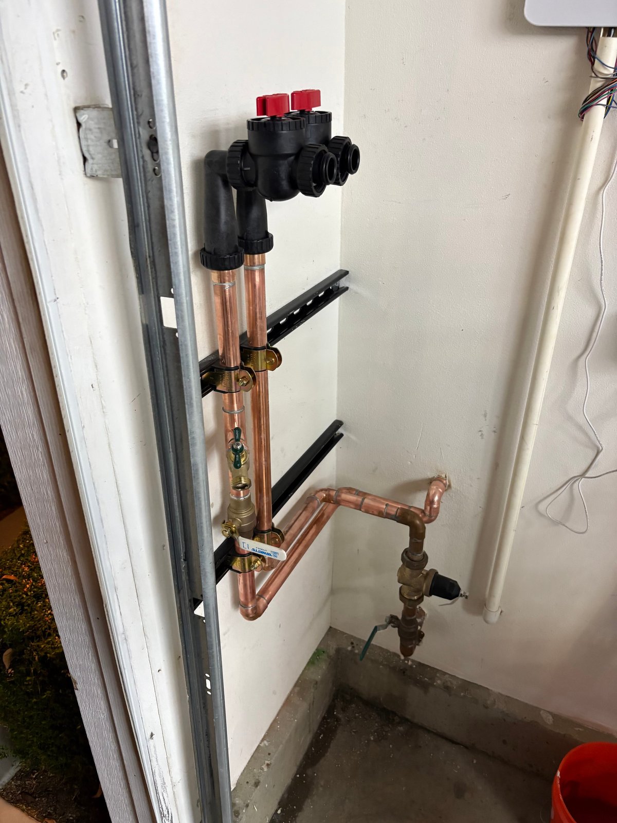 Plumbing manifold assembly with copper tubing, black shut-off valves, and red handles mounted on a white wall
